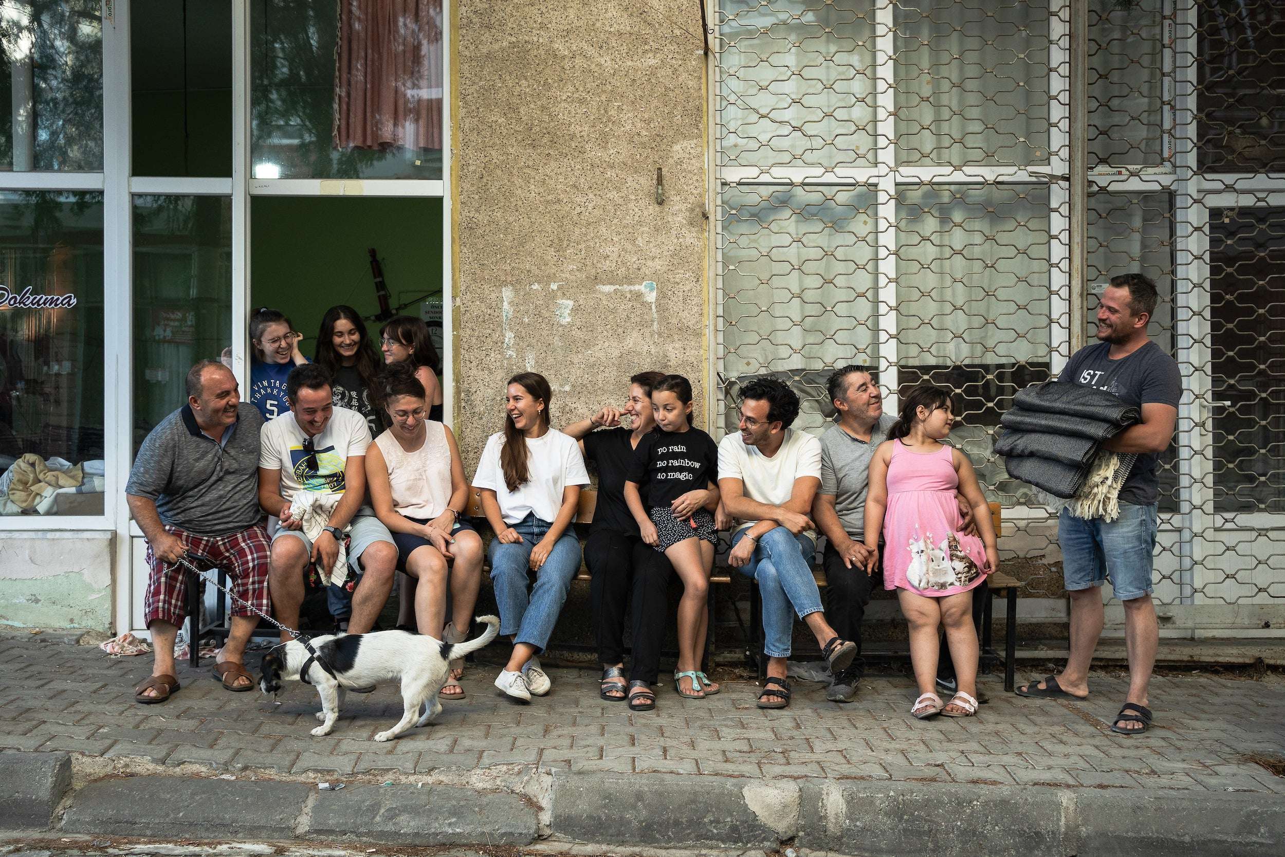 Dreizehn Menschen verschiedener Altersgruppen sitzen und stehen lachend vor einem Laden auf einer Bank, daneben läuft ein Hund